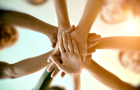 A group of individuals stack their hands together in a gesture of teamwork and unity, viewed from below against a bright, backlit backdrop.